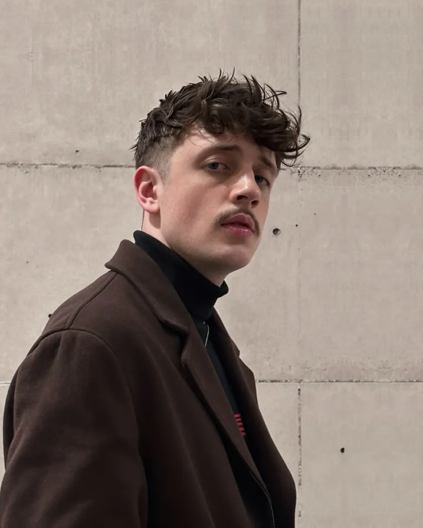 Isaac Eastley in a brown coat and black roll neck, standing against a concrete wall